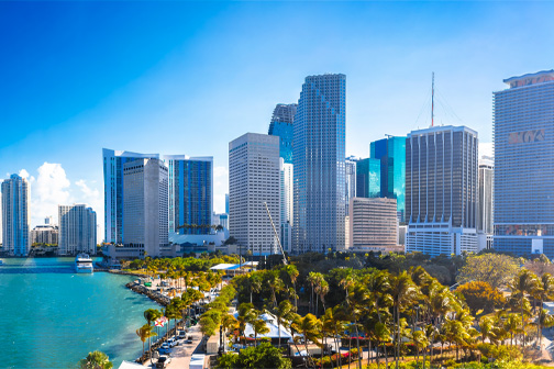Uitzicht op Miami met hoge gebouwen langs de kust, palmbomen en helderblauw water onder een zonnige lucht.